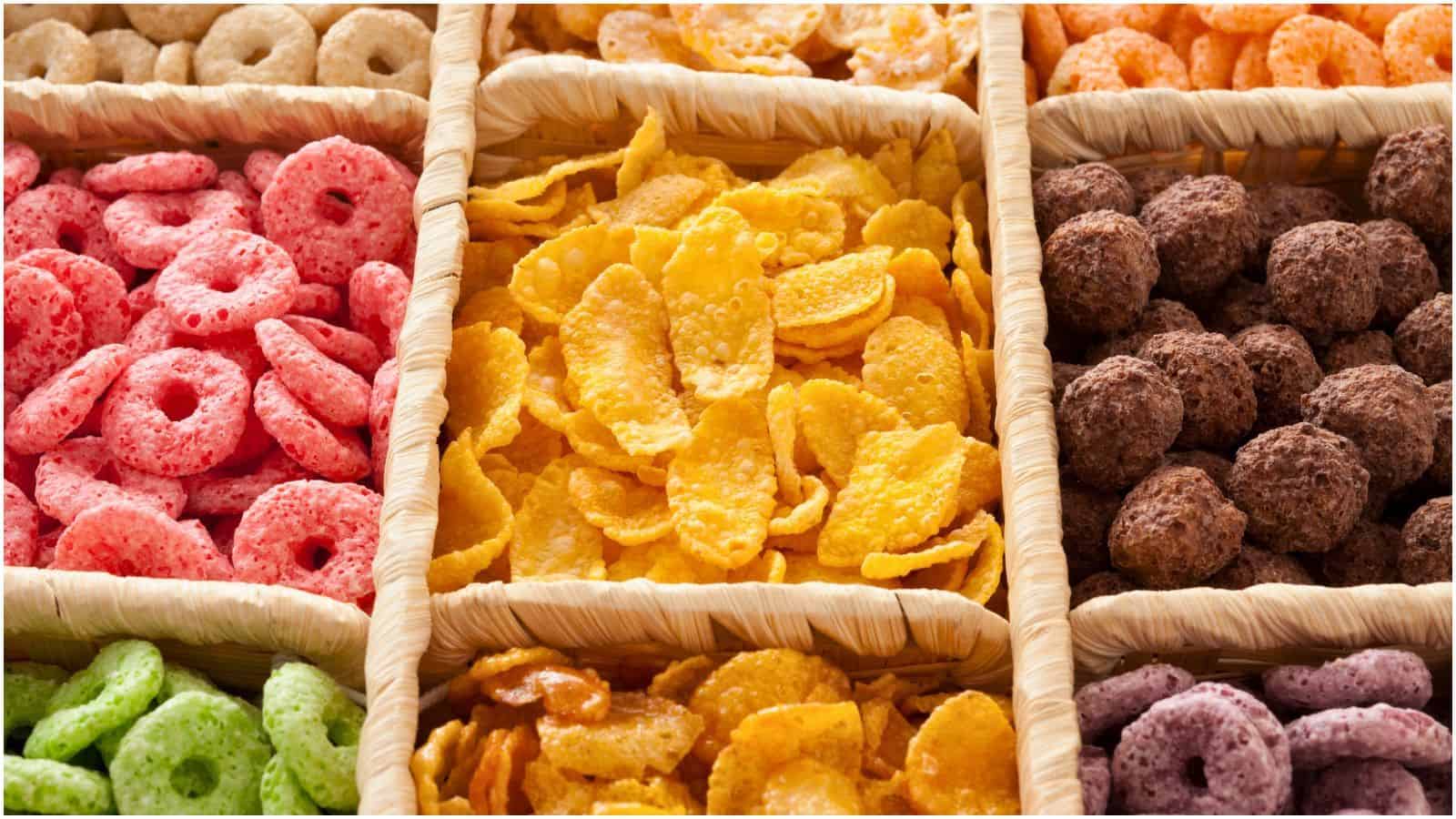 Rectangular baskets hold an assortment of colorful breakfast cereals. Varieties include pink, green, yellow, chocolate, orange, and purple cereals, each in their own section. The cereals are ring and ball shaped, with some flakes visible as well.