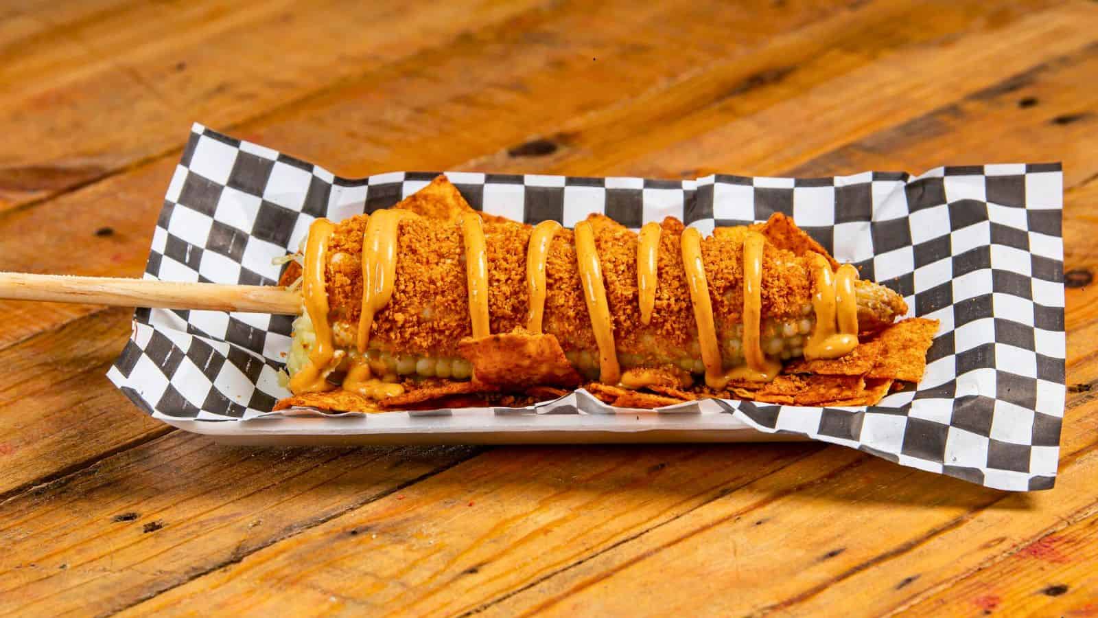 A piece of corndog drizzled in cheese sauce placed on a checkered plate