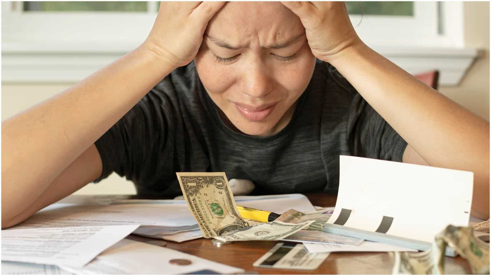 A person holds their head in frustration while looking at scattered bills, paperwork, and some cash on a table. The person appears stressed, with eyes closed and brow furrowed, suggesting financial troubles.