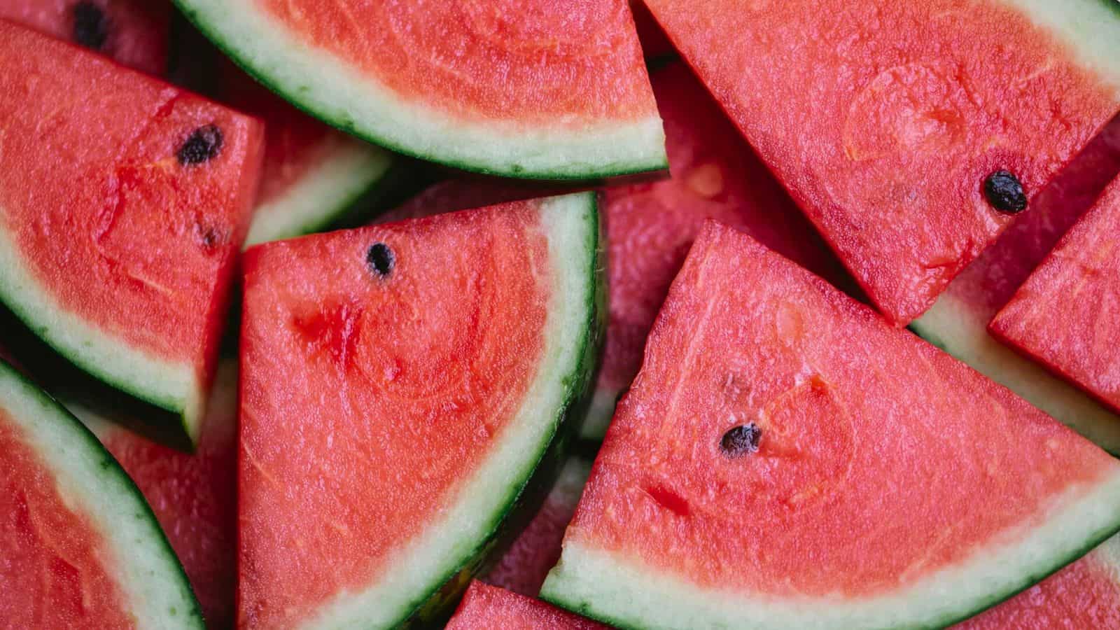 A close-up image shows several triangular slices of fresh watermelon. The watermelon slices display the bright red flesh, black seeds, and green rinds. The angled arrangement creates a visually textured and colorful composition.