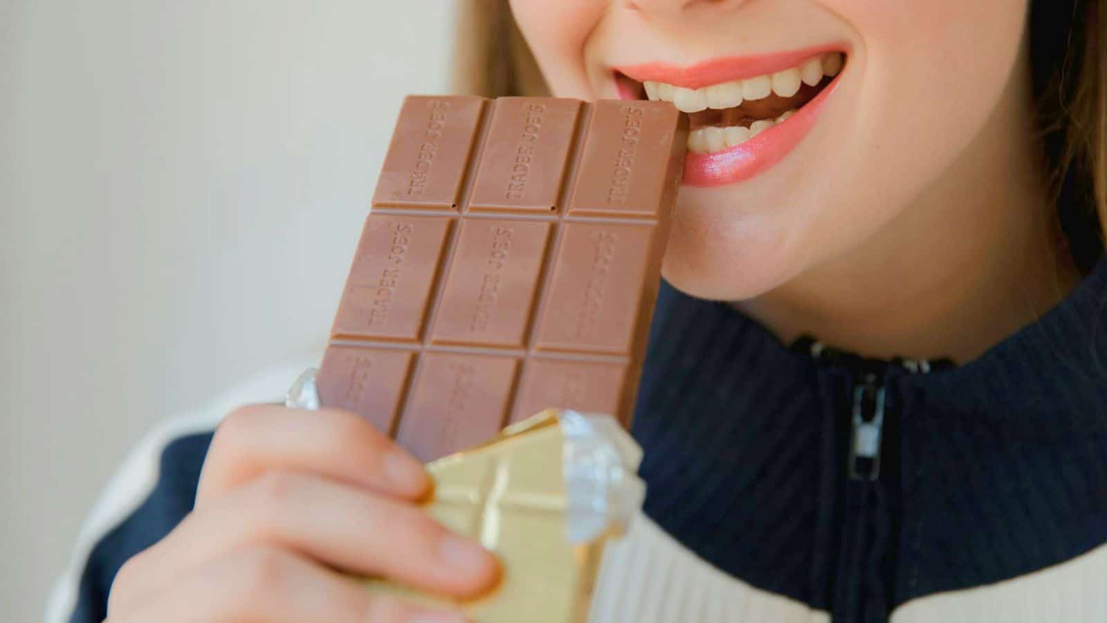 A person wearing a striped jacket is shown close-up taking a bite from a partially unwrapped chocolate bar. The person's smile is visible, and they appear to enjoy the chocolate.
