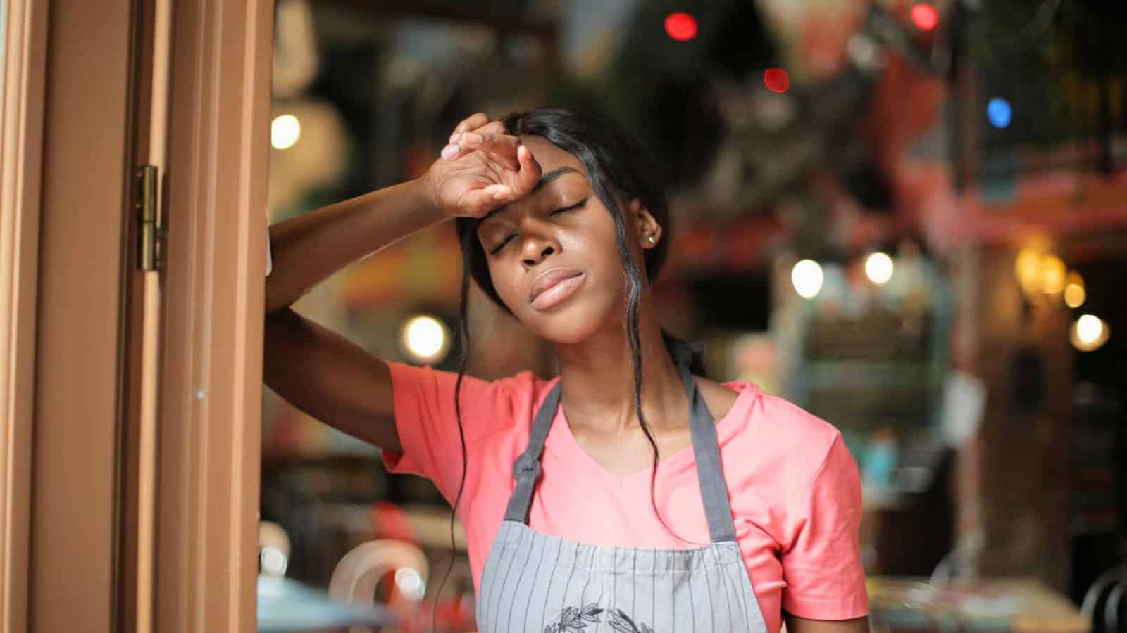 A person wearing a pink shirt and gray apron leans against a doorframe with their eyes closed. They appear to be tired, with one hand resting on their forehead. The background is a brightly lit interior space with colorful lights and decorations.