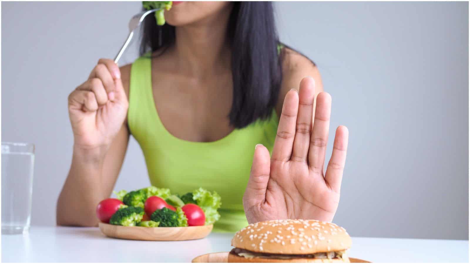 A person in a green tank top is seated at a table with a glass of water and a bowl of vegetables. They are eating a forkful of broccoli while holding up a hand to refuse a sandwich with a sesame seed bun placed on a plate in front of them.