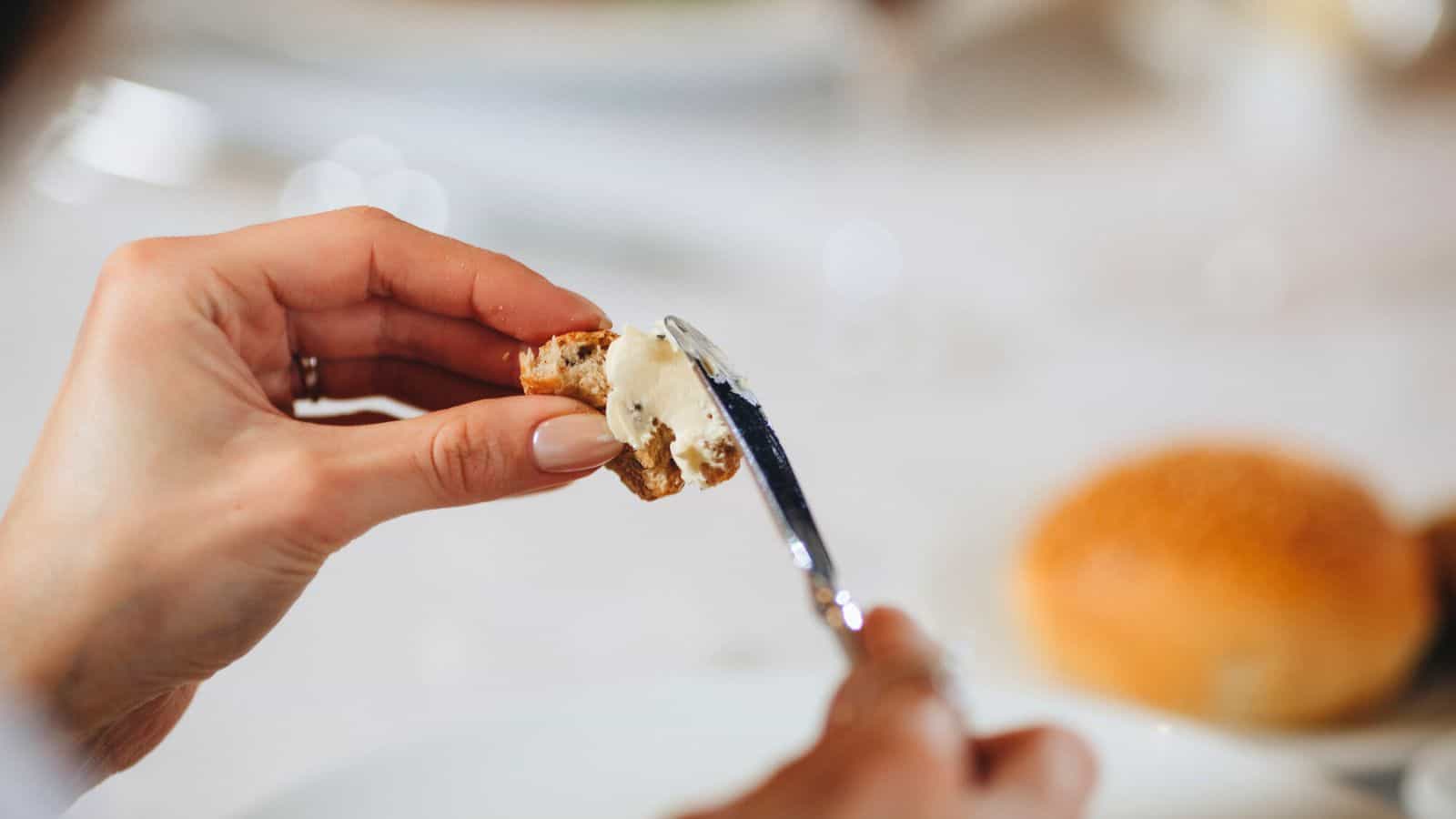 A close-up of a person spreading butter onto a piece of bread with a knife. A circular bread roll is visible in the blurred background on a white-covered table. The person's fingernails are painted in a light color.