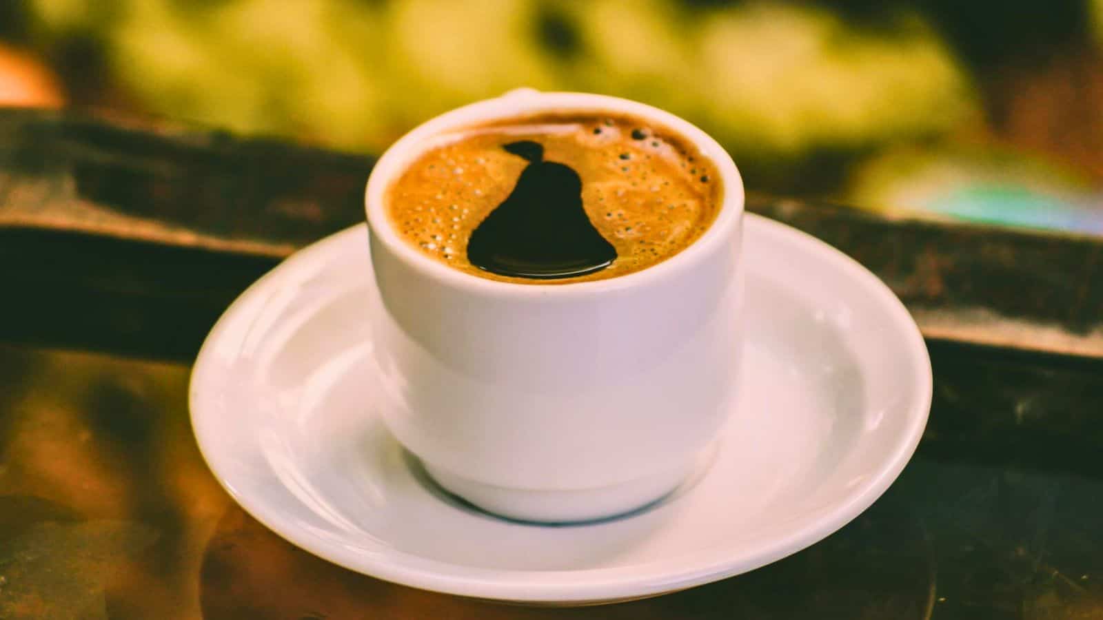 A white ceramic cup filled with dark coffee sits on a matching white saucer. The coffee has a distinctive shape on its surface, likely due to a splash effect. The background is out of focus with hints of green and brown tones.