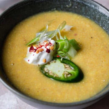 A bowl of yellow corn soup, reminiscent of famous dishes from the 60s, garnished with a dollop of sour cream, sliced green jalapeños, chopped green onions, and red chili flakes is shown. The bowl is placed on a light-colored surface. Additional sliced jalapeños are visible in a small dish nearby.