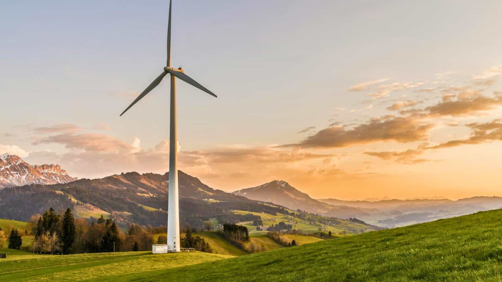 A landscape scene featuring a large wind turbine set in a grassy field. The background consists of rolling hills and distant mountains under a sky with scattered clouds during sunset. Trees and patches of forest can be seen near the turbine and on the hills.