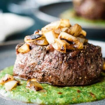 A close-up of a plated dish featuring a grilled steak topped with sautéed onions, sitting on a green sauce. The steak is garnished with herbs and the plate has a gray, minimalist design.