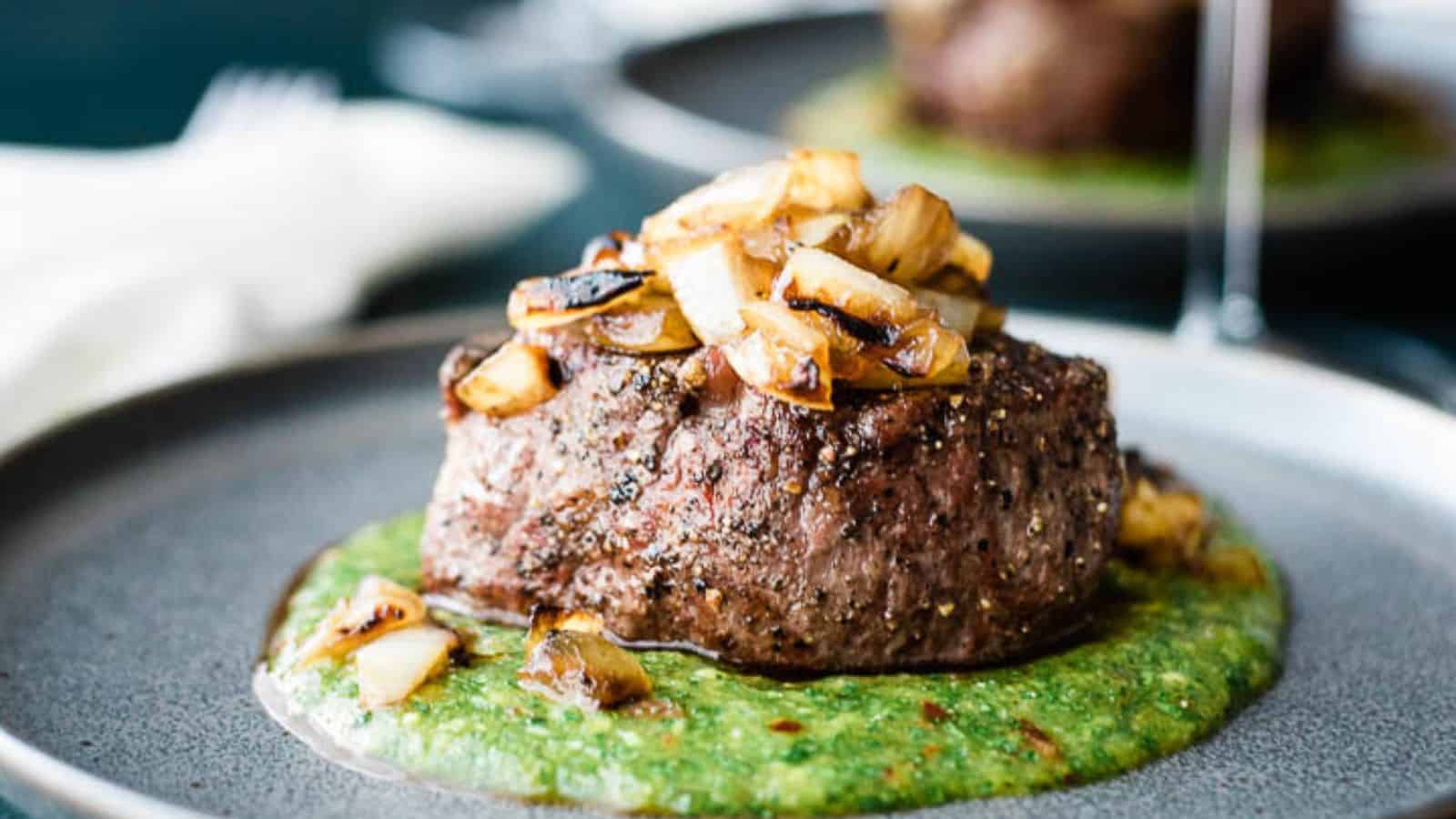 A close-up of a plated dish featuring a grilled steak topped with sautéed onions, sitting on a green sauce. The steak is garnished with herbs and the plate has a gray, minimalist design.