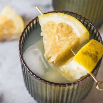 A close-up of a glass filled with an iced beverage, garnished with a lemon wedge on a cocktail pick. Another lemon wedge and part of a glass are visible in the blurred background, evoking the charm of iconic Sunday brunch drinks. The scene is set on a gray surface.