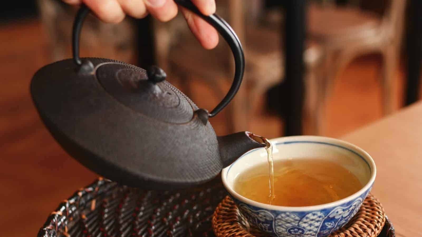 A hand pours tea from a black cast-iron teapot into a blue and white patterned ceramic cup placed on a wooden mat. The setting appears to be in a cozy indoor environment with chairs and tables in the background.