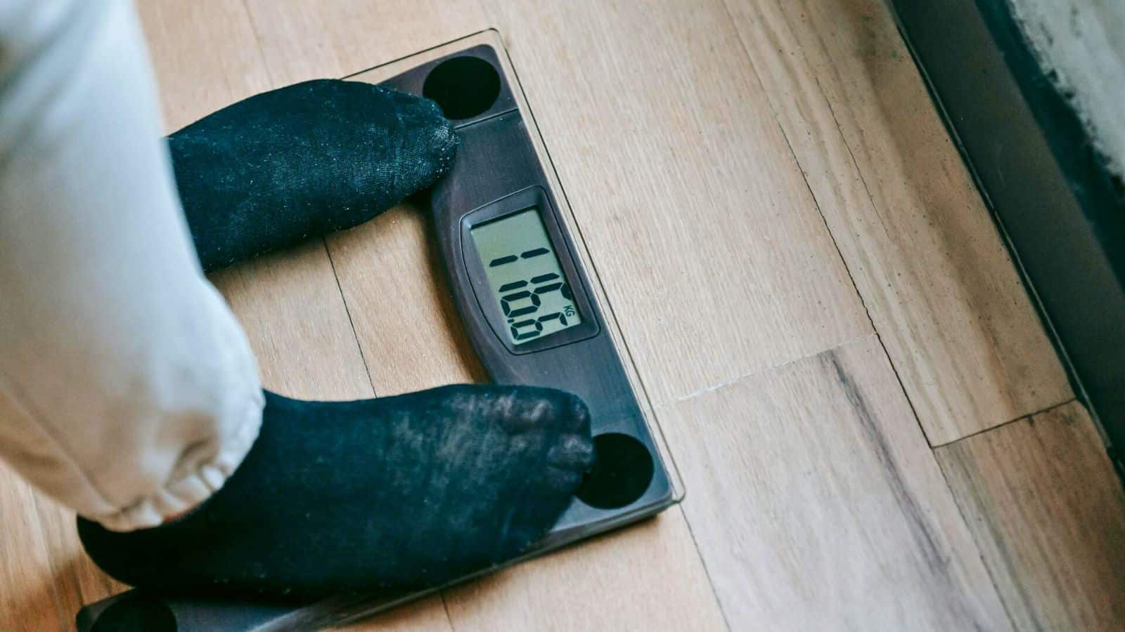 A person wearing black socks stands on a digital bathroom scale placed on a wooden floor. The scale's display shows a weight of 108.7 lbs. The person's feet are visible, and the image is taken from a top-down perspective.