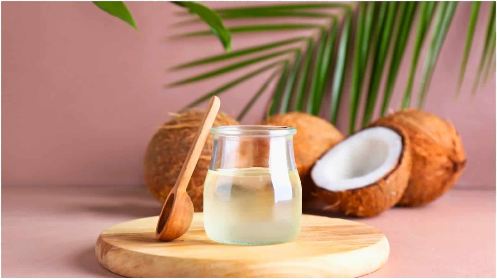 A glass jar of coconut oil with a wooden spoon rests on a round wooden board. In the background, there are three coconuts, one of which is split open, revealing the white flesh inside. Green palm fronds are partially visible against a pink backdrop.
