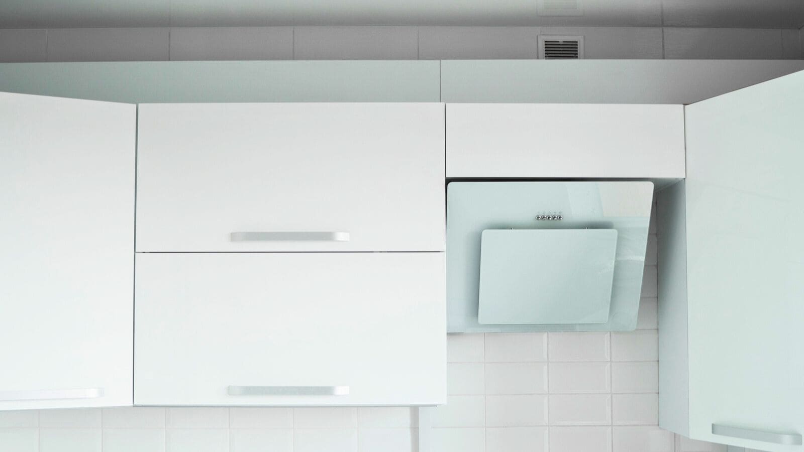 A modern kitchen with white cabinets and a sleek white range hood. The cabinets have horizontal handles, and the wall is tiled with white tiles, creating a clean and minimalistic aesthetic.