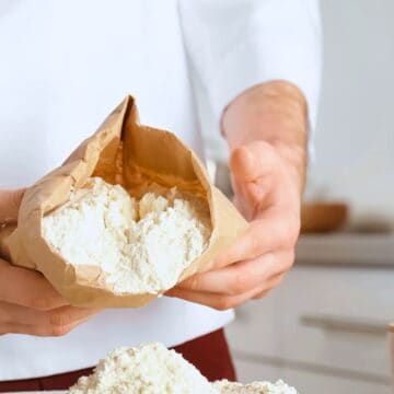 A person in a white chef's coat holding an open brown paper bag of powder, with some powder spilling out onto a kitchen counter. Various kitchen items, including a measuring cup and a small container, are partially visible in the background.