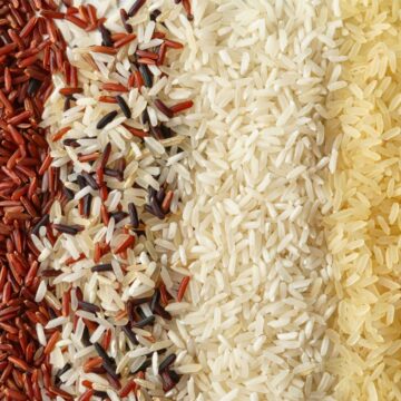A variety of rice grains arranged in vertical lines. From left to right, there are different types of rice: light brown, dark red, red with white, white with red, white, beige, and black rice. The grains showcase different sizes and textures.