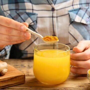 A person prepares a glass of ginger juice by adding a spoonful of turmeric powder. Fresh ginger roots are on a cutting board to the side, and there is a small bowl of turmeric powder next to the glass on a wooden surface. The person wears a checkered shirt.
