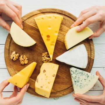 Four hands holding different types of cheese placed on a round wooden plate
