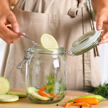 A person in a beige apron is placing a slice of vegetable into an open glass jar using a fork. Various sliced vegetables and herbs are on the wooden surface nearby, along with other jars in the background.