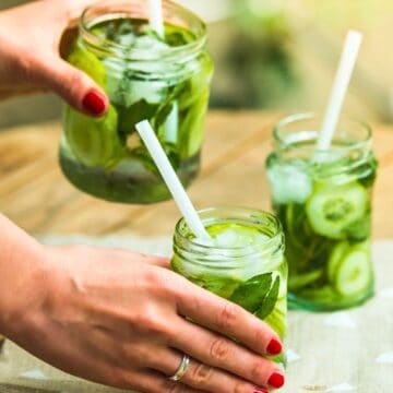 A person with red nail polish and a ring on their finger is placing a jar of a green drink with a white straw on a wooden table. Two other jars with the same drink, garnished with mint leaves and cucumber slices, are already on the table.