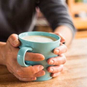 A person holding a light blue ceramic mug filled with a frothy beverage, such as coffee or hot chocolate. The person is sitting at a wooden table with hands wrapped around the mug. The background is slightly blurred.