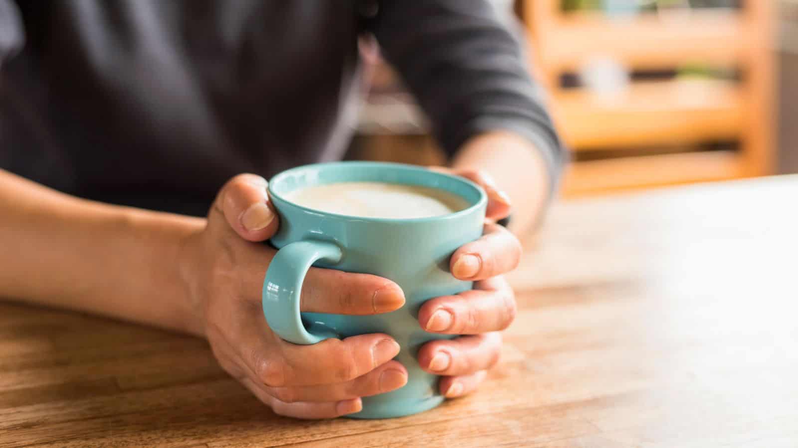 A person holding a light blue ceramic mug filled with a frothy beverage, such as coffee or hot chocolate. The person is sitting at a wooden table with hands wrapped around the mug. The background is slightly blurred.