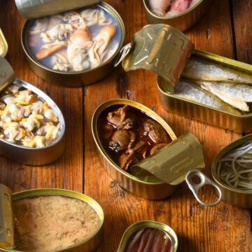 Open cans of various preserved seafood items are arranged on a wooden surface. They contain different types of fish, shellfish, cephalopods, and other preserved food items. Some cans have opened lids while others remain partially closed.