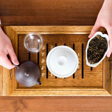 A person's hands are seen from above preparing tea. One hand is pouring from a small teapot into a teacup, while the other holds a dish with tea leaves. The setup includes a wooden tea tray with a small bowl and glass cup. The background is a wooden table.