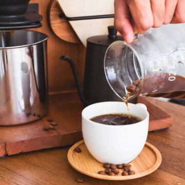 A person is pouring coffee from a glass carafe into a white cup placed on a wooden saucer. There is a coffee brewing setup with a stainless steel pitcher and a kettle on a wooden surface. Coffee beans are scattered on the table.
