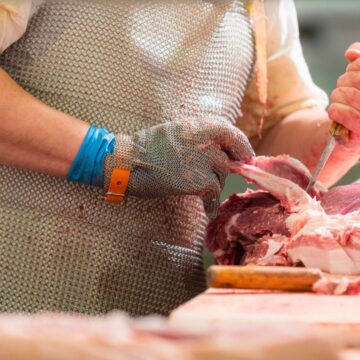 A butcher wearing a chainmail apron and protective gloves uses a knife to cut raw meat on a wooden cutting board. Plastic gloves are also visible over the chainmail gloves for additional hygiene.