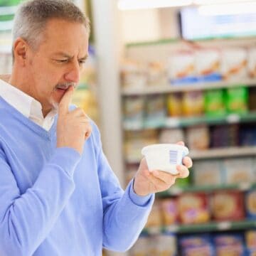 A man with a mustache, wearing a light blue sweater over a collared shirt, is standing in a grocery store aisle. He is looking intently at a container of food and holding it with one hand while touching his chin with the other. The shelves behind him are stocked with various products.
