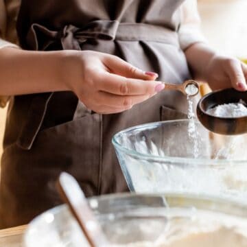 A person wearing a brown apron is adding baking powder from a teaspoon into a large mixing bowl filled with flour. Another mixing bowl is partially visible in the foreground. The scene is set in a well-lit kitchen.