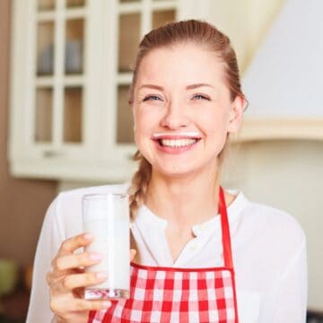 A woman with blonde hair, wearing a red and white checkered apron over a white shirt, smiles while holding a glass of milk. She stands in a kitchen setting with cabinets and kitchen items visible in the background, ready to share the health benefits of kefir.