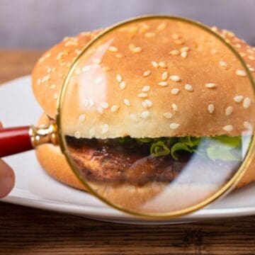 A hand holding a magnifying glass inspects a hamburger on a white plate. The hamburger has a sesame seed bun, lettuce, onions, and a visible patty. The background is a wooden surface.