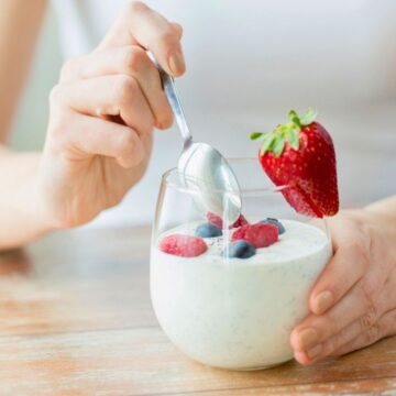 A person holding a bowl of yogurt topped with a strawberry, blueberries, and raspberries, and stirring it with a spoon. The person is wearing a white sleeveless shirt and is seated at a wooden table.