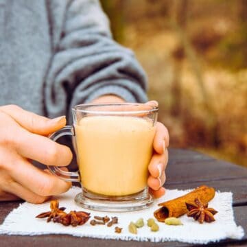 A person in a gray sweater holds a glass mug of chai tea with both hands. The mug sits on a white cloth on a wooden table. Surrounding the mug are cinnamon sticks, star anise, cardamom pods, and cloves. The background is blurred.