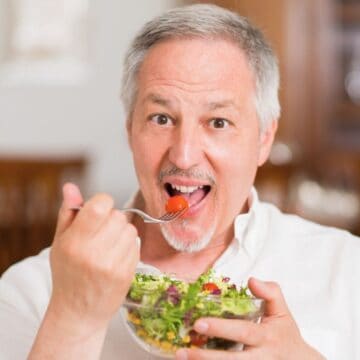 A middle-aged man with gray hair and a goatee is eating a salad out of a clear bowl. He is holding a fork with a cherry tomato in his mouth and is smiling. He is wearing a white shirt and sitting in an indoor setting with wooden furniture in the background.