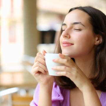 A person with long brown hair, wearing a purple top, sits indoors with their eyes closed and holds a white ceramic mug close to their face, appearing to enjoy the aroma. The background is blurred and shows light coming in through large windows.