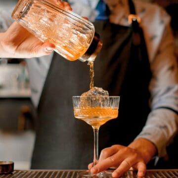 A bartender wearing a black apron pours a cocktail from a shaker into a glass with a large ice cube, standing at a bar with various bar tools and bottles blurred in the background.