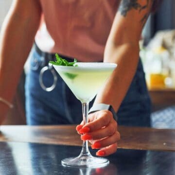 A person with a visible arm tattoo and painted fingernails is holding a green cocktail in a martini glass garnished with a mint sprig, placing it on a wooden table. Another person in the background is partially visible.