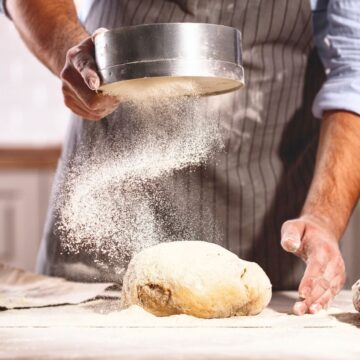 A person wearing a striped apron sifts flour onto a dough ball on a floured work surface with two loaves of bread nearby. The background features a blurred kitchen setting.
