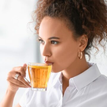A woman with curly hair and gold hoop earrings drinks a glass cup of tea while looking off to the side. She is wearing a white shirt and standing in a brightly lit room with a blurred background.