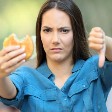 A woman with long brown hair wearing a blue shirt is holding a half-eaten hamburger in one hand and showing a thumbs-down with the other. She has a displeased facial expression, indicating dissatisfaction with the food. The background is blurred.