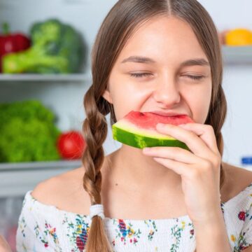 A young girl with braided hair is eating a slice of watermelon in a kitchen. She is holding another piece of watermelon in her left hand. The kitchen counter behind her has assorted vegetables, a loaf of bread, and containers of milk.