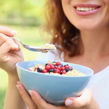 A person with wavy hair is smiling and holding a spoonful of cereal with berries and a blue bowl filled with the same. The background shows a blurry outdoor setting, possibly a park or garden, with greenery around.