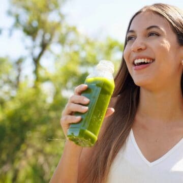 A woman with long brown hair holding a bottle of green juice while smiling. She is outdoors with trees and greenery in the background, wearing a white sleeveless top. The setting appears to be sunny, suggesting it's daytime.