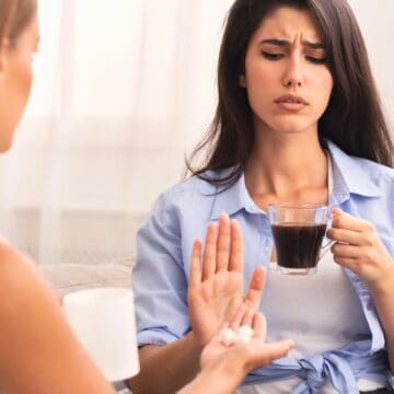 A woman with long dark hair holds a cup of coffee and raises her hand in refusal, looking displeased. Another person, whose face is not visible, extends an open hand offering pills. Both are seated indoors with a bright background.