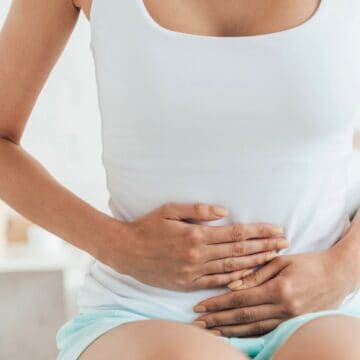 A person wearing a white tank top and light-colored shorts is sitting indoors on a bed, holding their stomach with both hands, suggesting discomfort or pain in the abdominal area. The background is blurred, focusing on the person.