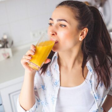 A woman in a kitchen drinks a glass of orange juice. She is wearing a white tank top with a blue and white patterned shirt over it. The kitchen has a sink, stove, and countertop with various items in the background.