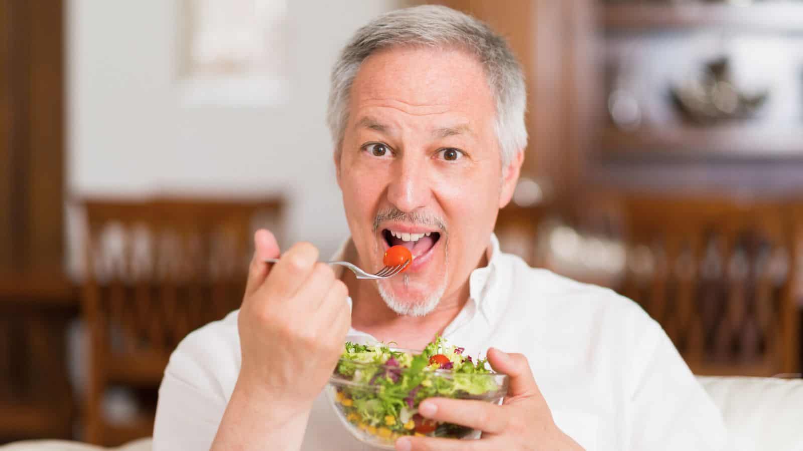 A middle-aged man with gray hair and a goatee is eating a salad out of a clear bowl. He is holding a fork with a cherry tomato in his mouth and is smiling. He is wearing a white shirt and sitting in an indoor setting with wooden furniture in the background.